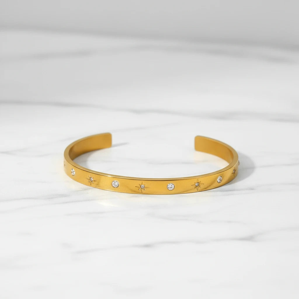 Minimalist product shot of a gold star cuff bracelet lying open on a white marble surface, the scattered star and crystal pattern giving it a celestial feel | Oria Jewel