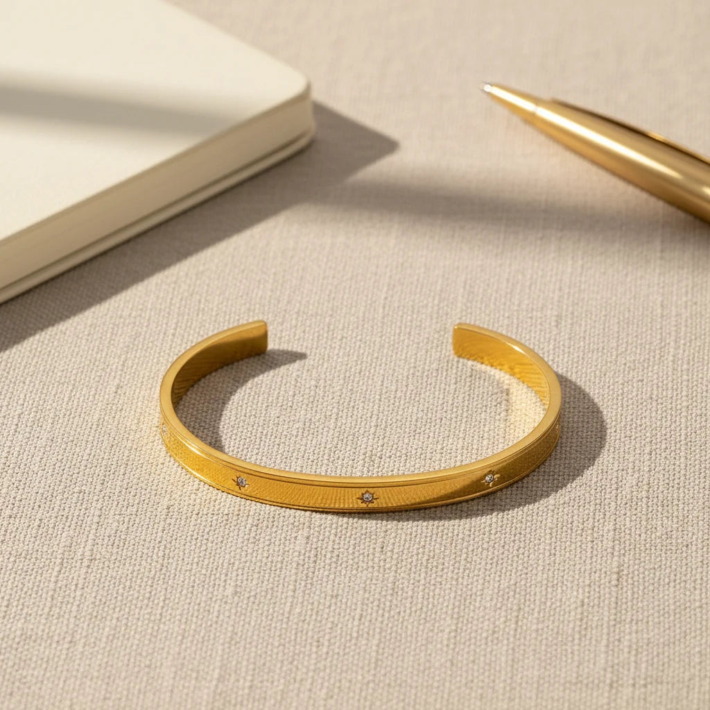 Flat-lay of a gold statement cuff bracelet on textured beige fabric next to a cream notebook and metallic pen, styled as a chic desk-side accessory | Oria Jewel