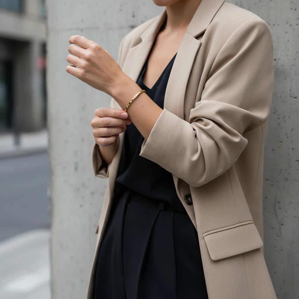 Woman in a beige blazer and black outfit adjusting a thin gold cuff bracelet on her wrist, demonstrating how the slim bangle adds a subtle, sophisticated touch to office wear | Oria Jewel