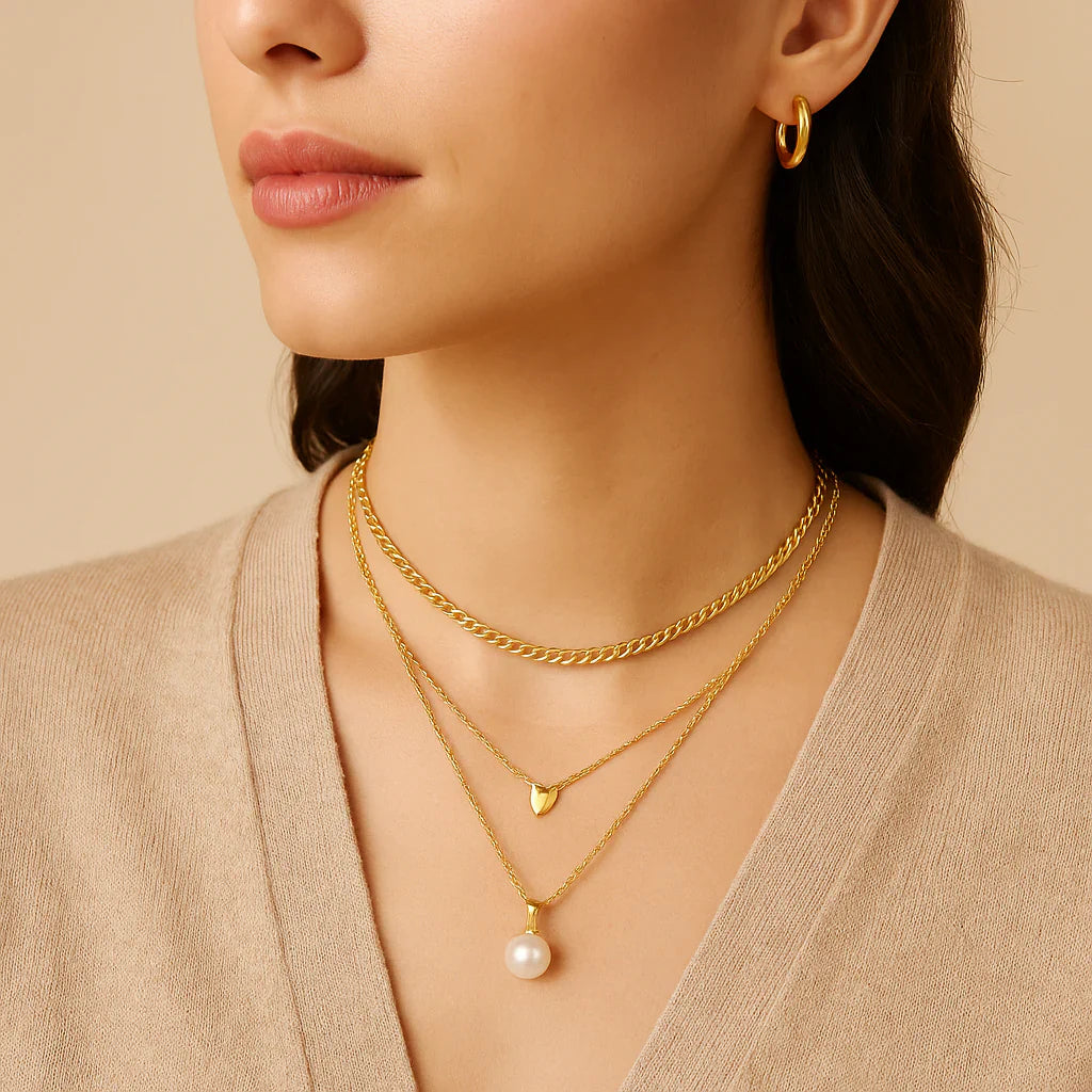Woman wearing layered gold necklaces—a chunky chain, a small heart pendant and a pearl drop—showing how to layer necklace styles with a simple beige V-neck top.