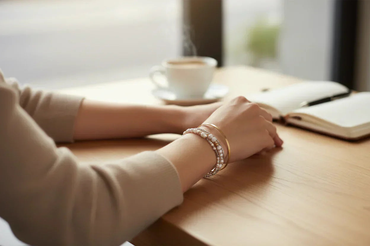 Woman resting her arms on a café table, wearing a simple stack of gold and beaded bracelets beside a coffee cup and notebook | Oria Jewel