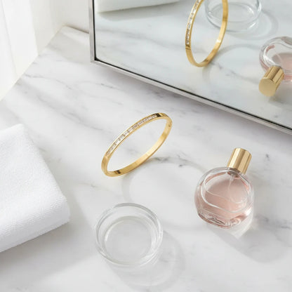 Flat lay on a marble vanity with mirror, perfume bottle, towel, and a sleek 14k gold hinged bangle bracelet placed in the center, highlighting its slim profile and stone detailing | Oria Jewel
