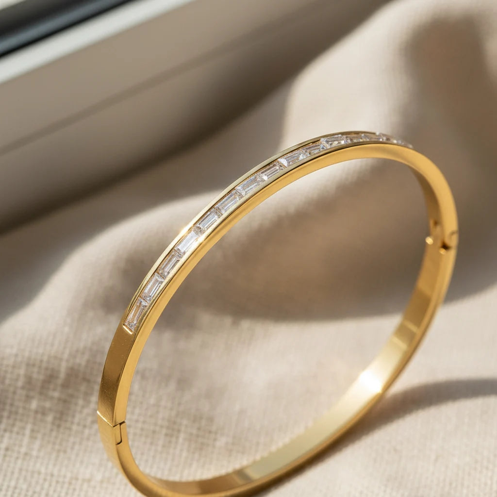 Close-up of a 14k gold hinged bangle bracelet resting on soft beige fabric near a window, focusing on the channel-set baguette stones along the top of the bracelet | Oria Jewel