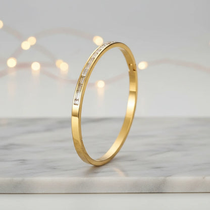 Standing hero shot of a 14k gold hinged bangle bracelet on a marble surface with soft fairy lights in the background, emphasizing its minimalist gold band and linear stones | Oria Jewel