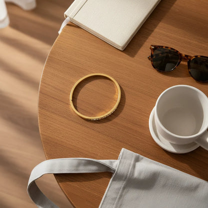 Top-down view of a wooden table with sunglasses, a white mug, and tote bag, featuring a 14k gold hinged bangle bracelet laid in a circle as the central accessory | Oria Jewel