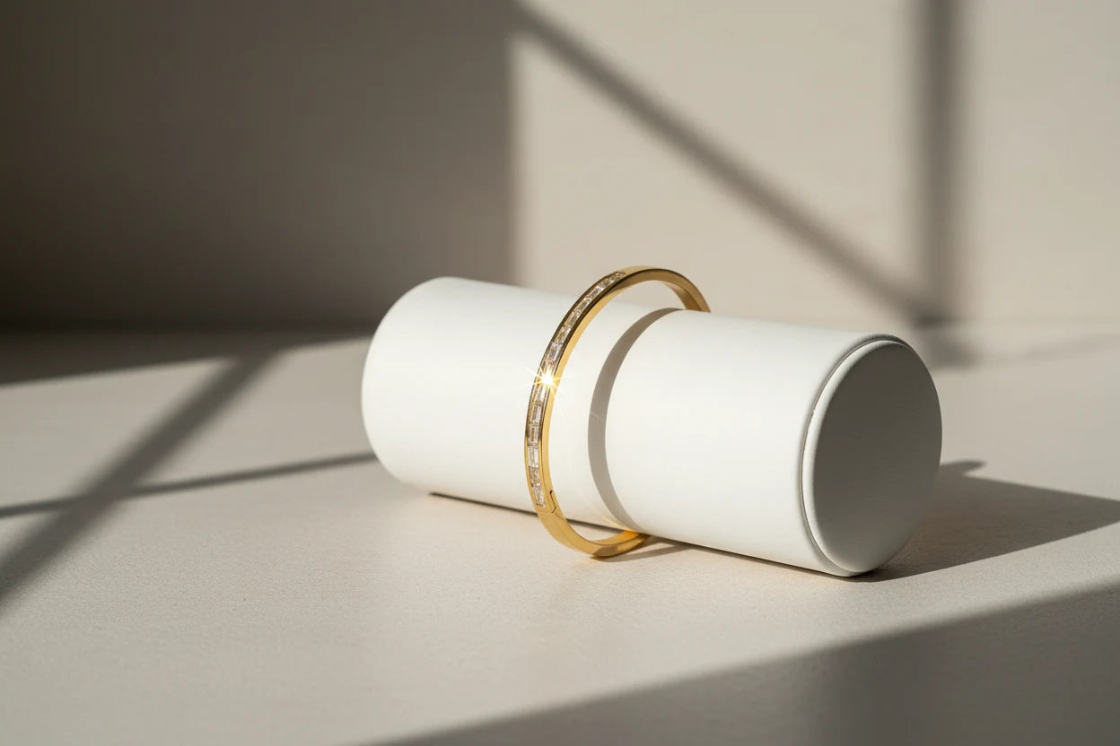 Hero image of a 14k gold hinged bangle bracelet balanced over a white cylindrical display in strong natural light, highlighting its glossy finish and slim baguette stone row | Oria Jewel