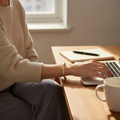 Woman working on a laptop at a wooden desk in warm daylight, with a fine 14k gold hinged bangle bracelet on her wrist adding a polished touch to her casual sweater and trousers | Oria Jewel