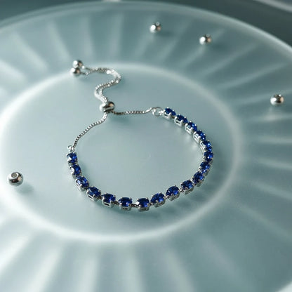 Silver Aurelia Cluster Tennis Bracelet with vivid deep-blue round crystals arranged in a neat row, styled on a frosted glass surface with soft radial light reflections and scattered metal beads for a modern, luminous look | Oria Jewel