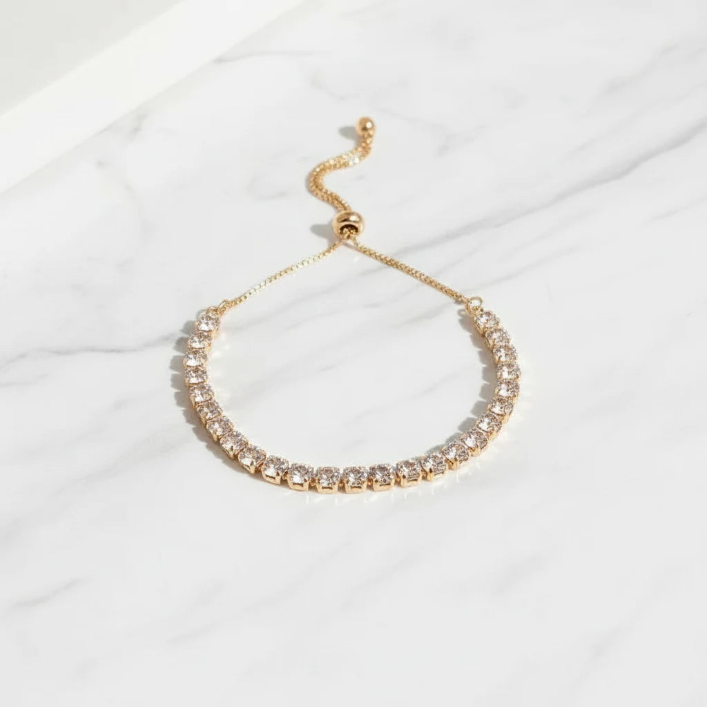 Elegant gold Aurelia Cluster Tennis Bracelet displayed in a soft curve on white marble, catching natural light to highlight each clustered crystal stone | Oria Jewel