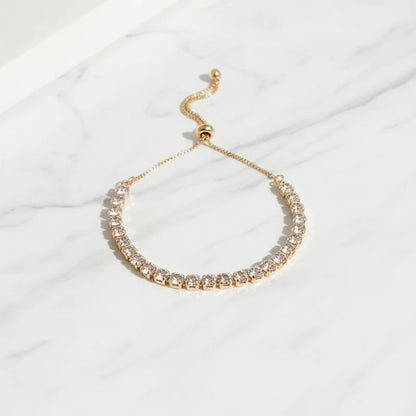 Elegant gold Aurelia Cluster Tennis Bracelet displayed in a soft curve on white marble, catching natural light to highlight each clustered crystal stone | Oria Jewel