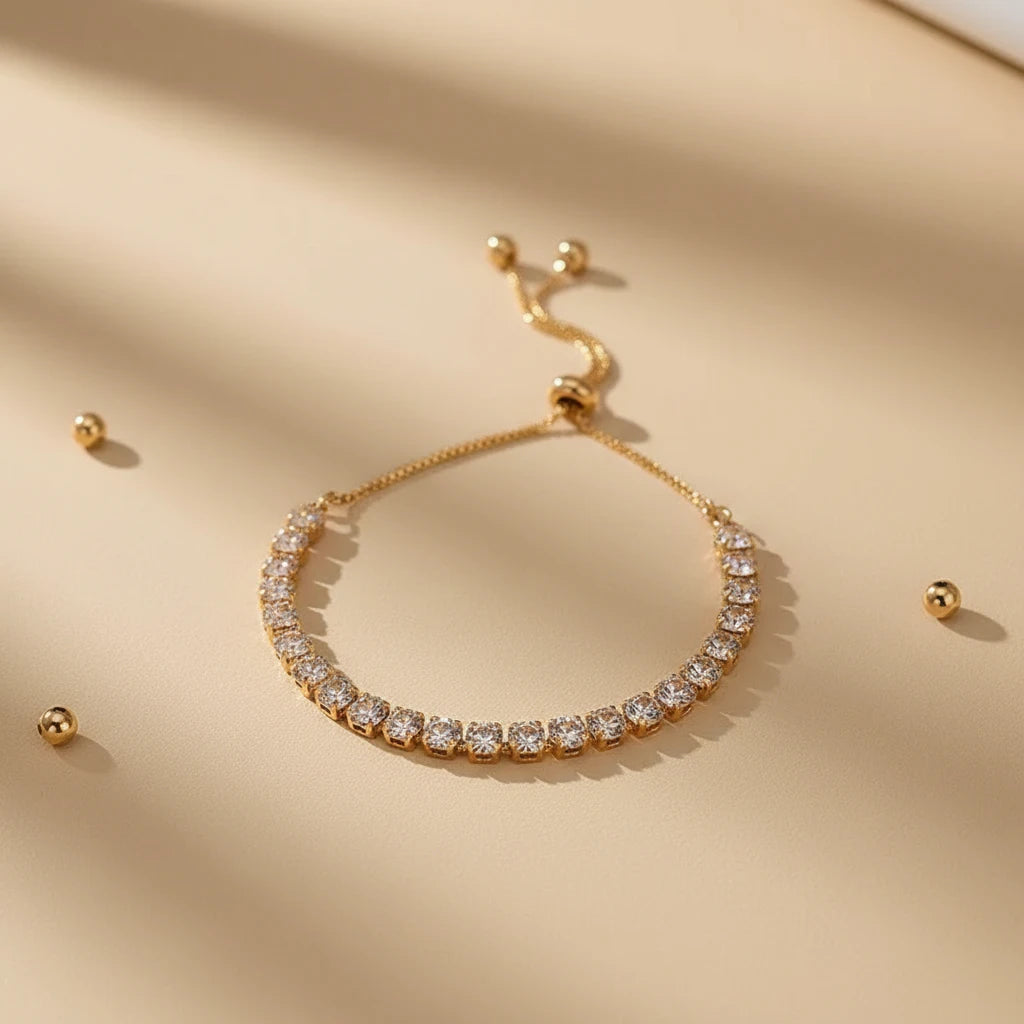 Gold Aurelia Cluster Tennis Bracelet styled on a beige surface with scattered metallic beads and soft shadows, highlighting the dainty adjustable chain and crystal row | Oria Jewel