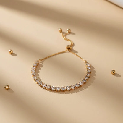 Gold Aurelia Cluster Tennis Bracelet styled on a beige surface with scattered metallic beads and soft shadows, highlighting the dainty adjustable chain and crystal row | Oria Jewel