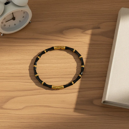 Black Bamboo bangle Bracelet with gold details resting on a wooden desk beside a white alarm clock and fabric notebook in warm sunlight | Oria Jewel