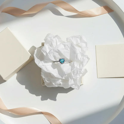 Blue Heart Halo CZ Ring styled as a gift flat-lay with ribbon and crinkled tissue on a white tray, featuring a bright blue heart-cut center stone framed by a sparkling halo on a pavé band | OriaJewel
