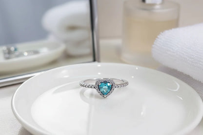 Blue Heart Halo CZ Ring close-up on a glossy white dish with a mirror and perfume bottle softly blurred behind, highlighting the blue heart gemstone, halo CZ border, and pavé-set silver band | OriaJewel