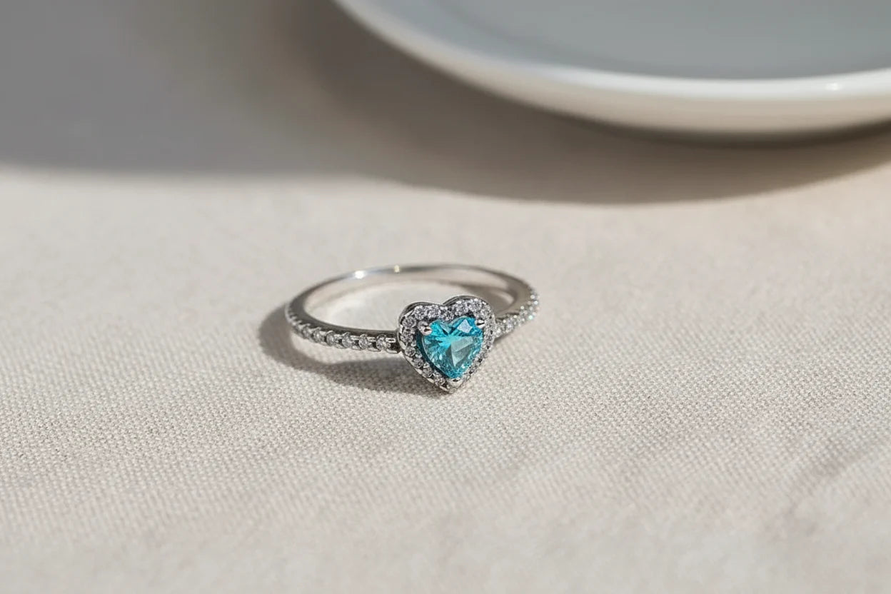 Blue Heart Halo CZ Ring macro on textured beige fabric near a white plate, natural sunlight catching the halo sparkle around the blue heart center and the pavé stones along the band | OriaJewel