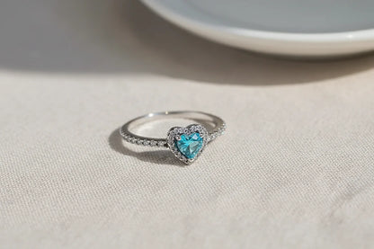 Blue Heart Halo CZ Ring macro on textured beige fabric near a white plate, natural sunlight catching the halo sparkle around the blue heart center and the pavé stones along the band | OriaJewel