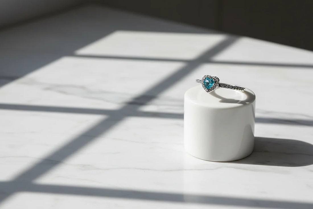 Blue Heart Halo CZ Ring displayed on a white cylindrical stand under window-grid shadows on marble, showcasing the heart-shaped blue stone, halo setting, and slim pavé band | OriaJewel