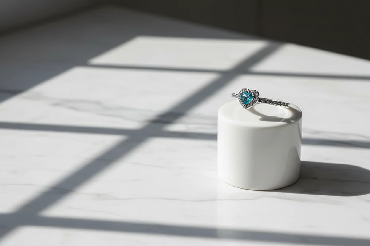 Blue Heart Halo CZ Ring displayed on a white cylindrical stand under window-grid shadows on marble, showcasing the heart-shaped blue stone, halo setting, and slim pavé band | OriaJewel