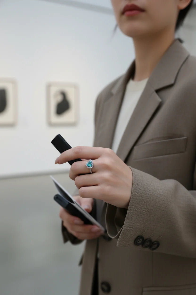 Blue Heart Halo CZ Ring worn with a blazer outfit in a gallery-style setting while holding a phone and notebook, focus on the blue heart center stone and shimmering halo CZ frame | OriaJewel