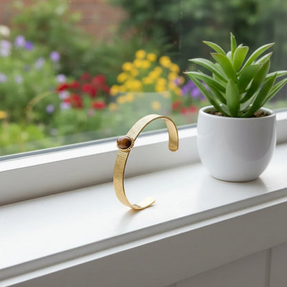 Brown gemstone cuff bracelet standing upright on a bright windowsill next to a small white planter with a green succulent and blurred colorful garden in the background | Oria Jewel