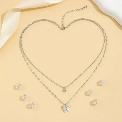 Flat lay of a silver Butterfly Layered Necklace arranged in a heart shape on a beige background with scattered crystal beads and ribbon, highlighting its dainty sparkle | Oria Jewel