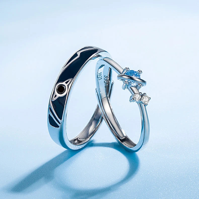 Cosmic Heart Couple Ring set on a cool blue background, showing the matching silver rings—one with black enamel cosmic pattern and one with a blue heart stone plus small clear accents—styled in a clean studio shot | OriaJewel