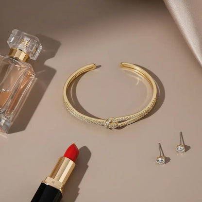 Flat lay of a gold Crystal Knot Cuff Bracelet beside perfume, red lipstick, and stud earrings on a beige background | Oria jewel 
