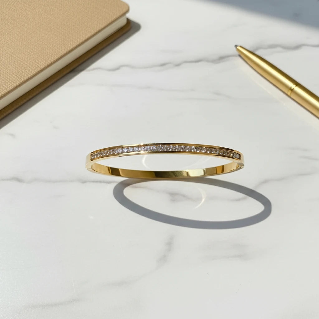 Crystal Line Gold Bangle laid on a white marble surface beside a beige notebook and gold pen, the bracelet’s linear crystal row and mirror-polished gold band reflecting soft daylight for a clean, minimal flat-lay composition | Oria Jewel
