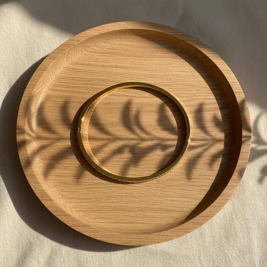 Simple flat-lay of a plain polished Crystal Line Gold Bangle resting inside a round wooden tray, warm sunlight casting leaf-shaped shadows that emphasize the smooth golden circle and minimalist aesthetic | Oria Jewel