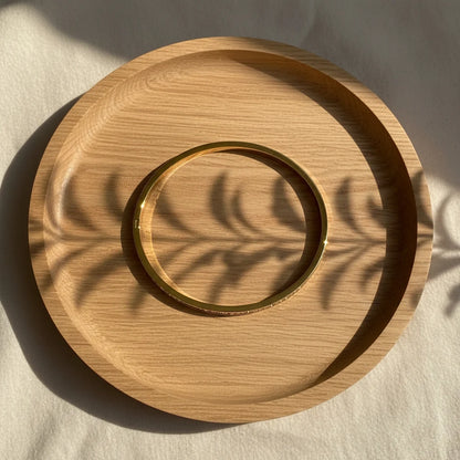 Simple flat-lay of a plain polished Crystal Line Gold Bangle resting inside a round wooden tray, warm sunlight casting leaf-shaped shadows that emphasize the smooth golden circle and minimalist aesthetic | Oria Jewel