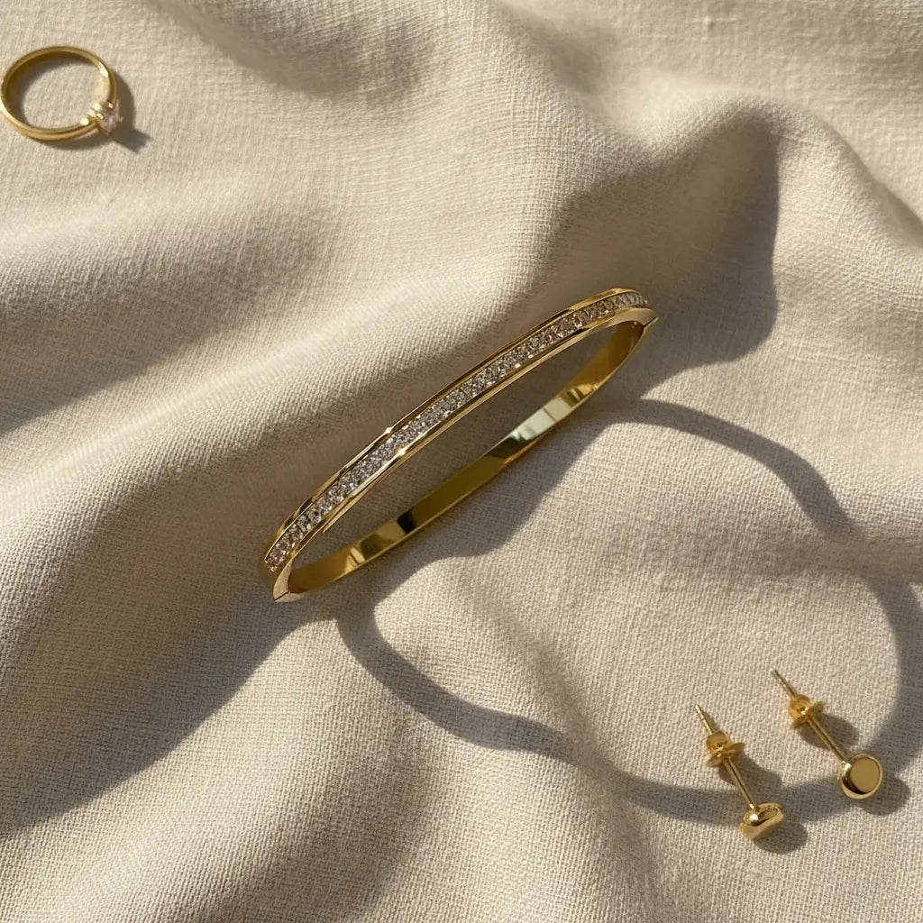 Crystal Line Gold Bangle styled on soft beige fabric next to a matching gold ring and stud earrings, the bracelet’s full line of crystals and shiny gold band creating a coordinated, sunlit jewelry set perfect for gifting | Oria Jewel