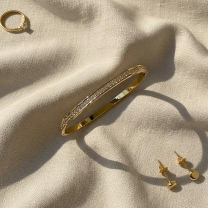 Crystal Line Gold Bangle styled on soft beige fabric next to a matching gold ring and stud earrings, the bracelet’s full line of crystals and shiny gold band creating a coordinated, sunlit jewelry set perfect for gifting | Oria Jewel