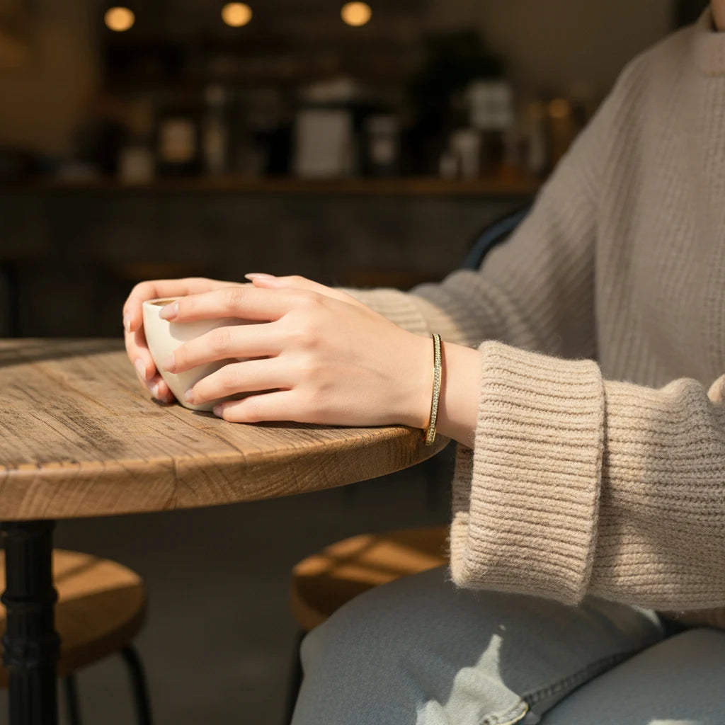 Cozy café moment with a woman in a chunky beige knit sweater holding a warm cup of coffee, her wrist adorned with the slim Crystal Line Gold Bangle that adds a subtle row of sparkle to the relaxed, everyday look | Oria Jewel