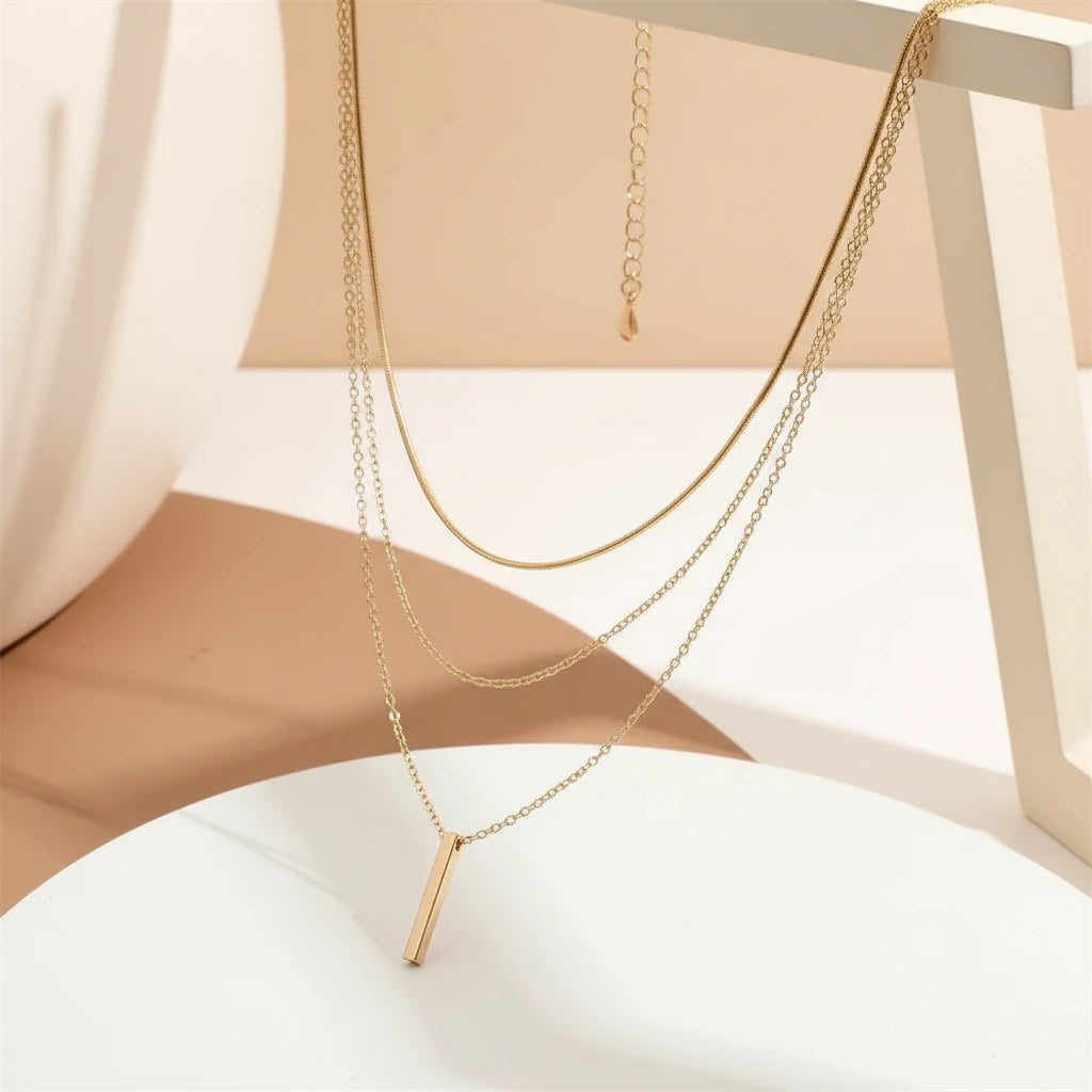 Drop Bar Pendant Necklace with triple gold chain hanging gracefully on modern geometric stand | Oria Jewel