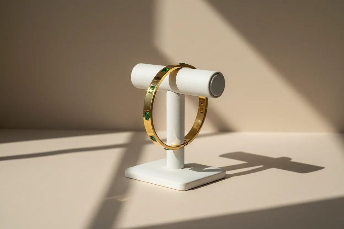 Emerald Gold Hinged Bangle displayed upright on a white T-bar stand, sunlight creating dramatic shadows and a star-like sparkle on one green stone against a soft beige backdrop | Oria Jewel