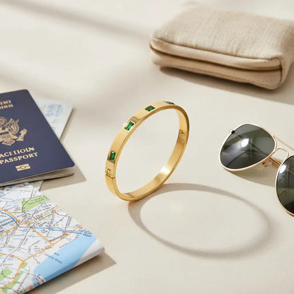 Flatlay of Emerald Gold Hinged Bangle styled for travel beside a folded map, U.S. passport, beige wallet, and gold-rimmed sunglasses on a neutral surface | Oria Jewel