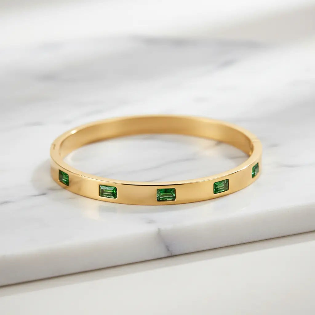 Close-up of Emerald Gold Hinged Bangle resting on white marble, showing a full row of rectangular green stones set around the polished gold band | Oria Jewel