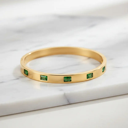 Close-up of Emerald Gold Hinged Bangle resting on white marble, showing a full row of rectangular green stones set around the polished gold band | Oria Jewel