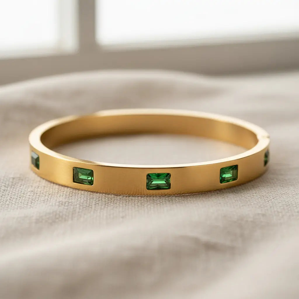 Emerald Gold Hinged Bangle on soft beige fabric near a window, matte gold finish and vivid green stones highlighted by natural daylight for a cozy, elegant feel | Oria Jewel