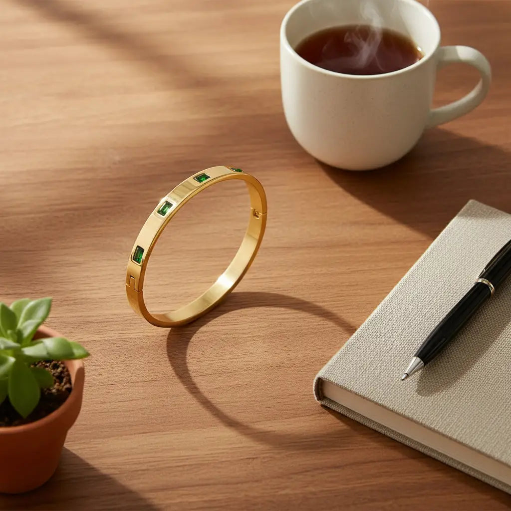 Emerald Gold Hinged Bangle standing on its edge on a wooden desk next to a steaming cup of tea, small plant, and notebook with black pen, creating a warm work-from-home scene | Oria Jewel