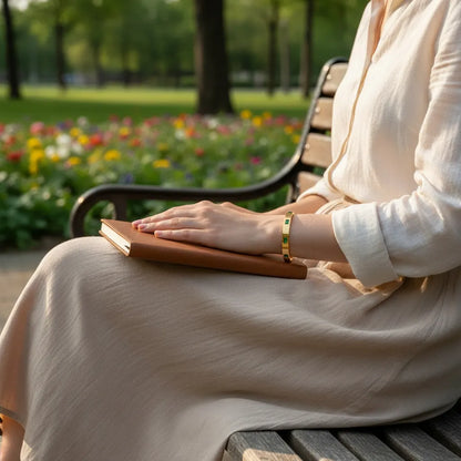 Woman sitting on a park bench in a flowing cream dress, hands resting on a brown notebook while wearing the Emerald Gold Hinged Bangle, with colorful flower beds blurred in the background | Oria Jewel