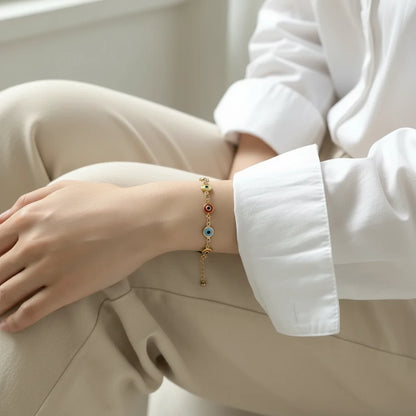Close-up of a woman sitting with folded legs by a bright window, wearing a delicate Evil Eye Charm Bracelet with multicolored enamel charms on a gold chain, styled with a crisp white shirt and beige trousers | Oria Jewel