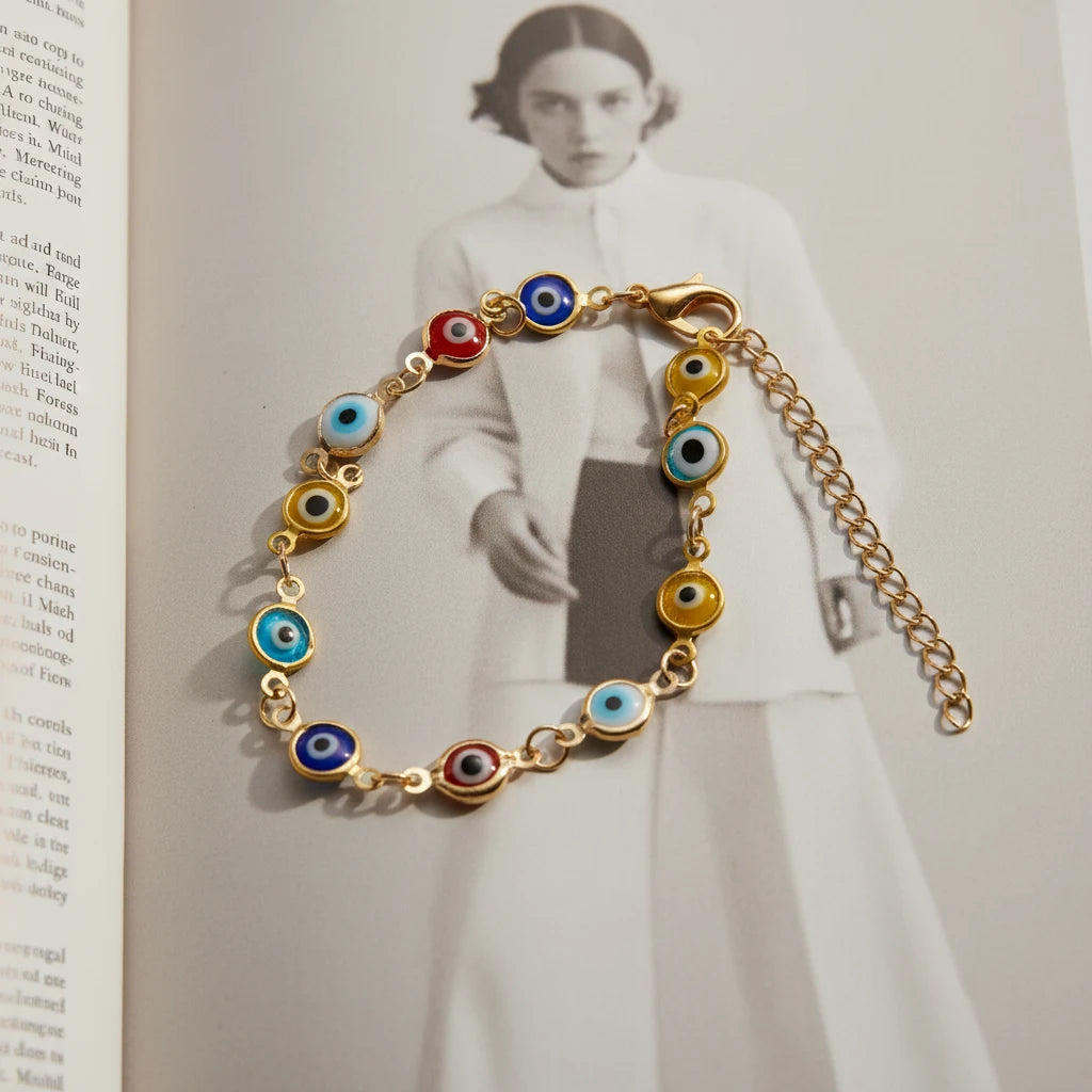 Editorial-style image of the Evil Eye Charm Bracelet laid out on an open fashion magazine, showcasing the vivid red, blue, yellow, and turquoise glass eye charms linked on a fine gold chain | Oria Jewel