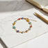 Overhead shot of the Evil Eye Charm Bracelet resting on a white marble surface beside a notebook and gold pen, the multicolored eye charms forming a neat circle that highlights its protective, modern design | Oria Jewel