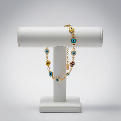 Studio display of the Evil Eye Charm Bracelet draped over a white jewelry stand, emphasizing the gold chain and alternating blue, yellow, red, and turquoise evil eye charms in a clean, minimalist setting | Oria Jewel