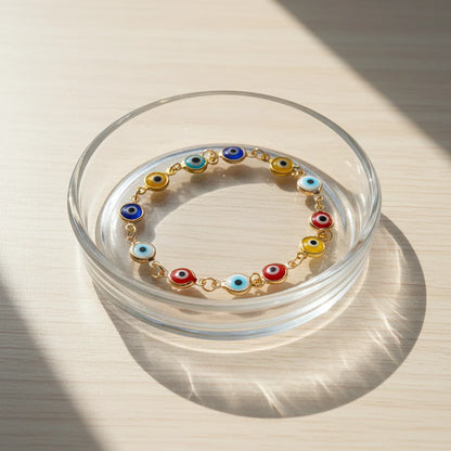 Sunlit close-up of the Evil Eye Charm Bracelet arranged inside a clear glass dish on a wooden surface, the colorful glass eye charms glowing softly in the natural light for a bright, uplifting vibe | Oria Jewel