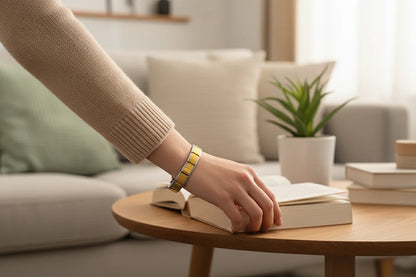 Cozy living room scene showing a woman’s arm reaching for a book on a round wooden coffee table, the Gold Center Stainless Steel Stretch Bracelets adding a warm pop of gold against her beige knit sweater | Oria Jewel