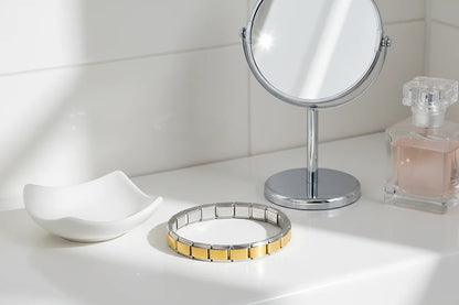 Bathroom vanity scene with Gold Center Stainless Steel Stretch Bracelets resting on a glossy white countertop next to a round mirror and perfume bottle, sunlight reflecting softly off the gold center links | Oria Jewel