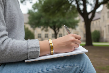 Outdoor lifestyle shot of a woman writing in a notebook on her lap, wearing Gold Center Stainless Steel Stretch Bracelets over a grey sweater, with a blurred campus background creating a relaxed, studious mood | Oria Jewel